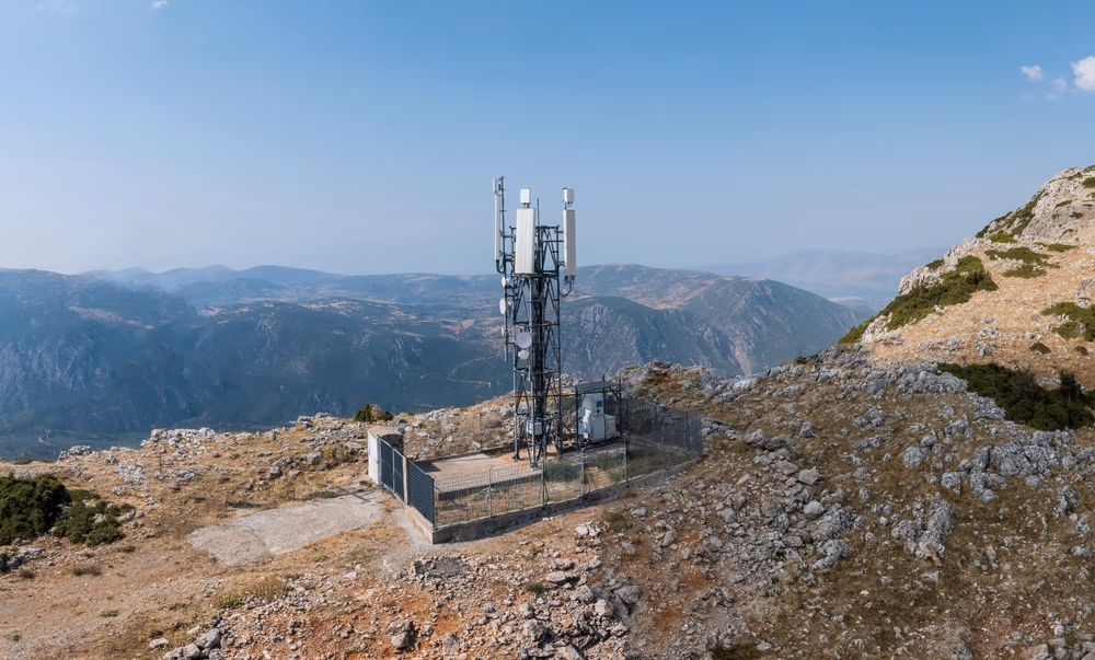 Cellular communication tower on a rocky mountain peak, equipped with Edge Technologies, is surrounded by a fenced enclosure; distant mountains and a clear sky stretch in the background.