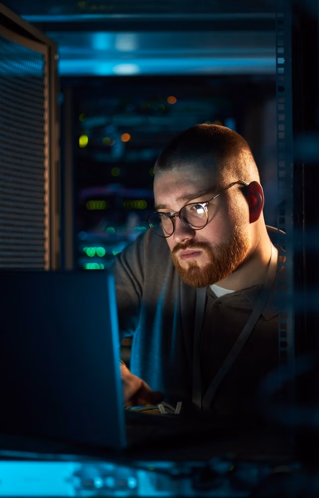 A man with glasses and a beard works on a laptop in a dimly lit server room, surrounded by server racks and electronic equipment, focusing on large-scale data processing.
